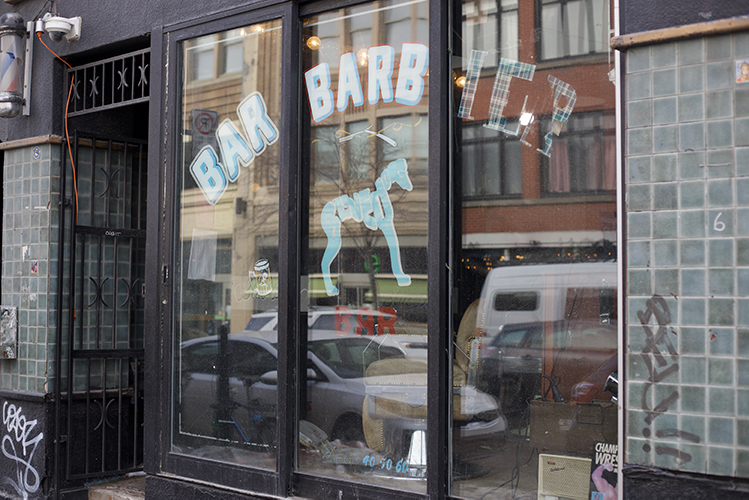 The outside of Blue Dog Bar and barbershop. The logo of a blue dog is pasted on the window, along with a sign that reads "Bar Barbier."