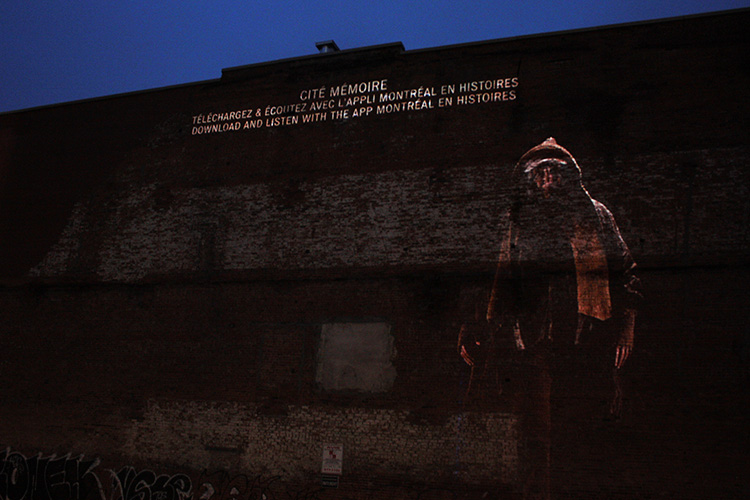 A projection of a hooded man on a plain brick wall.