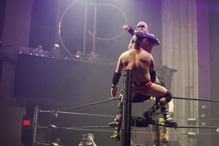In the corner of the ring, one wrestler grabs the hair of another, slightly above him, and winds up to hit him in the face.