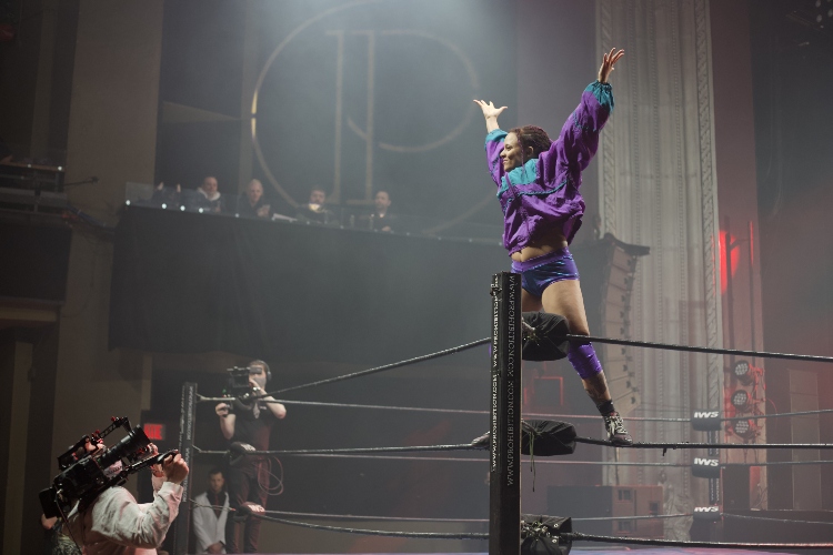A woman stands on the ropes in the corner of the ring and lifts her arms up and open. She cheers with the crowd.