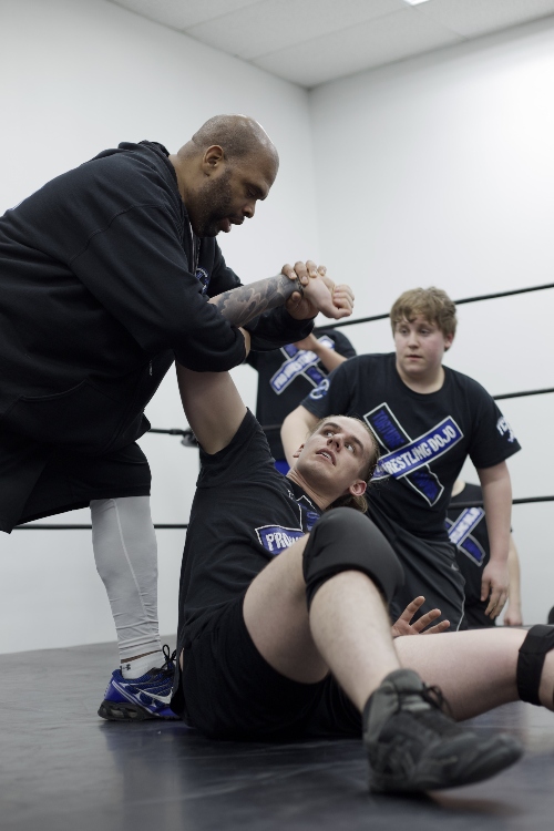 The pro wrestling coach grabs the arm of one of his students as other wrestlers watch from behind.