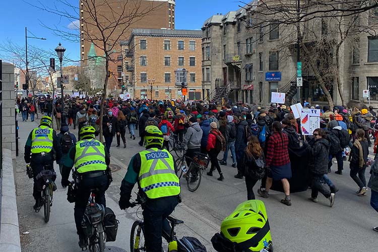 Many people are walking down a street to demonstrate. They are surrounded by police officers on bicycles.