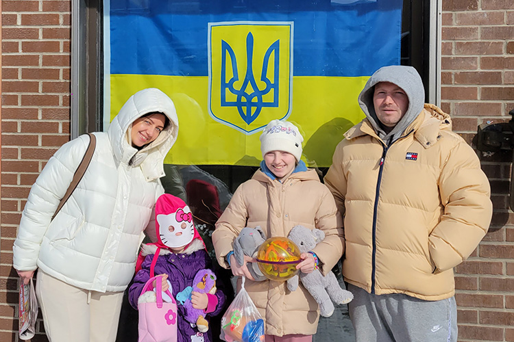 The family of four stands in front of a large blue and yellow Ukrainian flag.