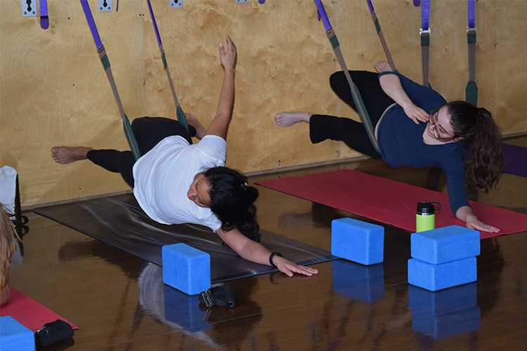 Two women hang from purple wall straps upholstered to the wall, with yoga matts on the floor. The straps are supporting their bodies in a side-lunge position.