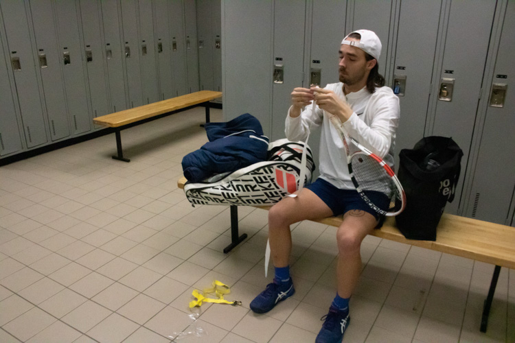 Alec Brideau wrapping his tennis racket with white grip in the locker room
