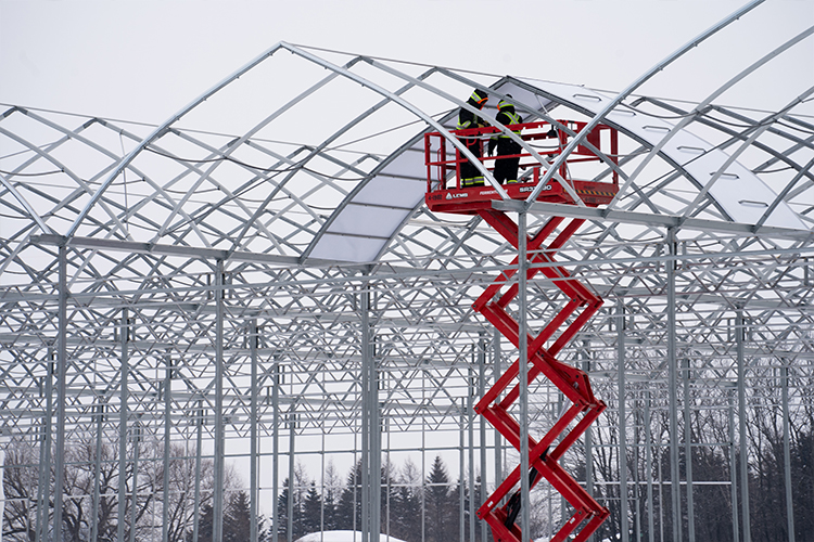 Construction workers on a red lift outside large metal vertical farm structures with a grey winter sky
