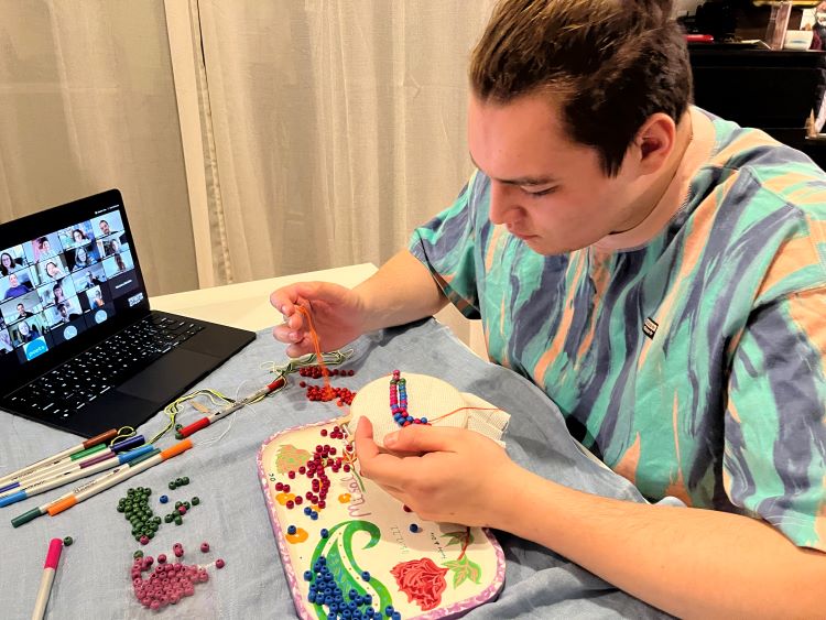 a student sowing beads into his embroidery project. His laptop is out in front of him showing a zoom call with various participants.