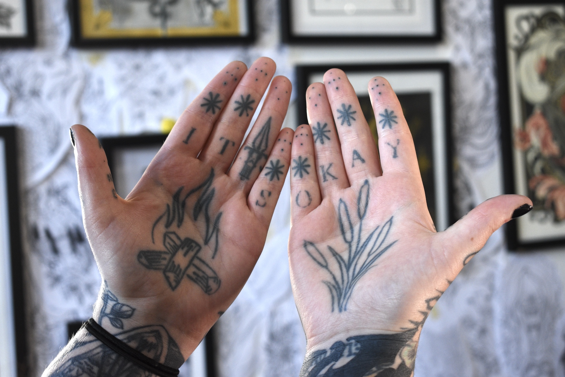 Hands side by side in front of wall full of art. Hand tattoos read "It's okay."