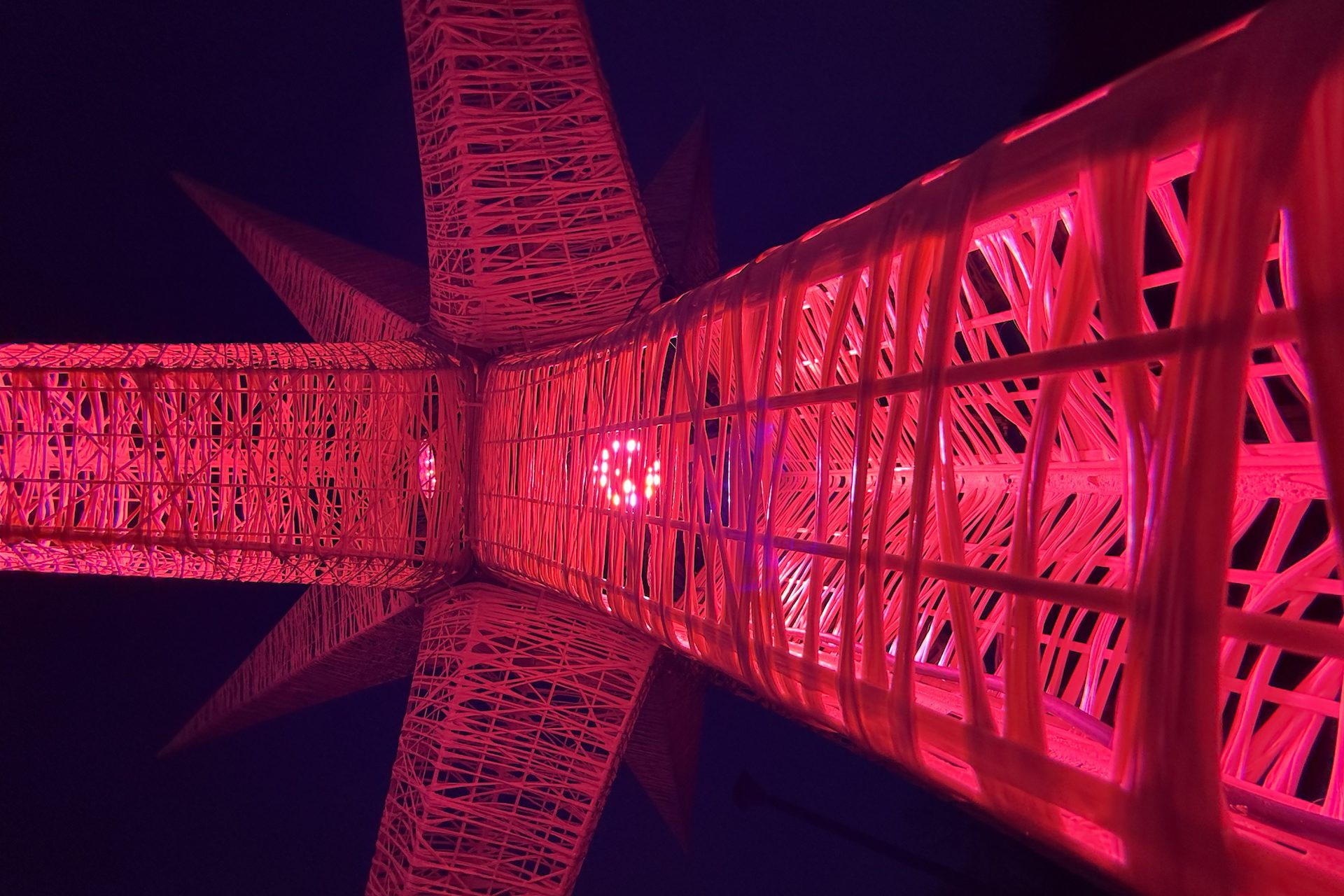 A public art structure, illuminated in red, stretches across a blue background